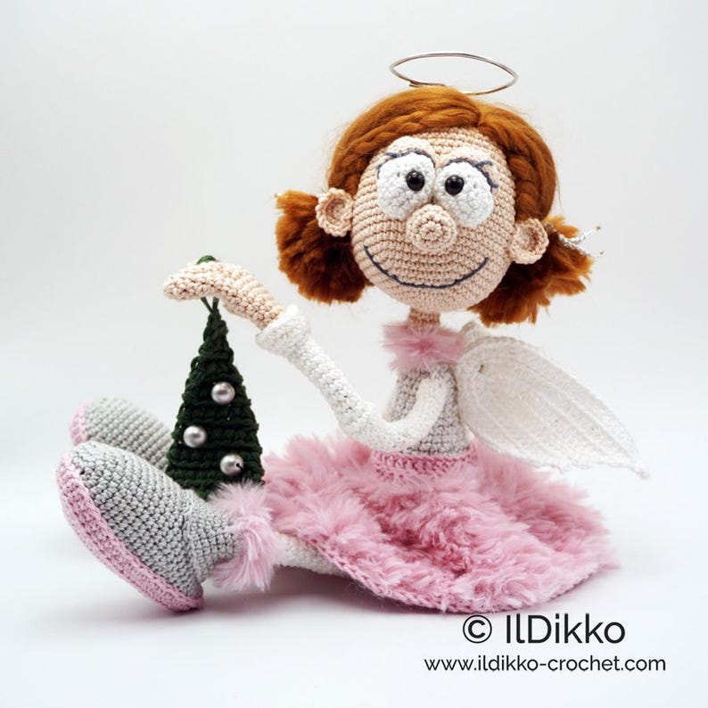 May include: A crocheted angel doll with auburn hair, a wire halo, and white wings. The doll wears a pink fuzzy skirt, white long sleeves, and gray boots with pink soles. It holds a small dark green crocheted Christmas tree with silver beads.