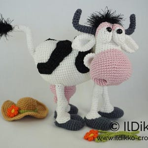 May include: A crocheted cow toy with black and white spots, pink ears, and a black tail. The cow is wearing a brown crocheted hat with an orange flower.