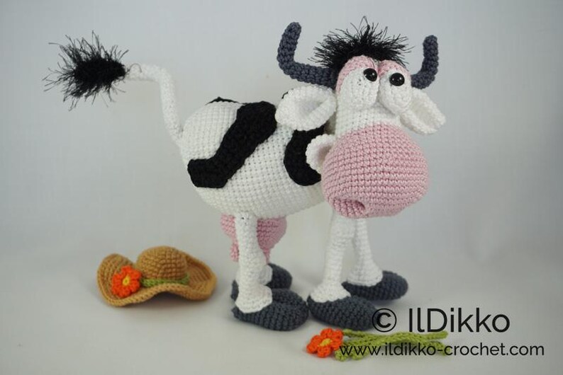 May include: A crocheted cow toy with black and white spots, pink ears, and a black tail. The cow is wearing a brown crocheted hat with an orange flower.