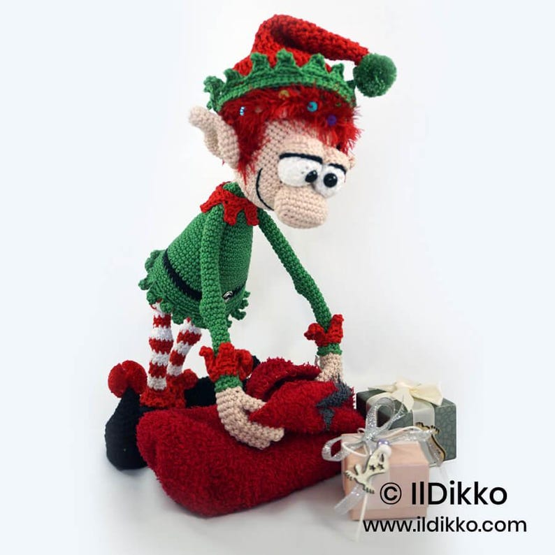 May include: A crocheted elf doll with red hair, a green tunic, and a red hat with a green pom-pom. The elf has large white eyes and is reaching towards a red fabric bundle. Two small gift boxes with ribbons are placed next to the elf figure.
