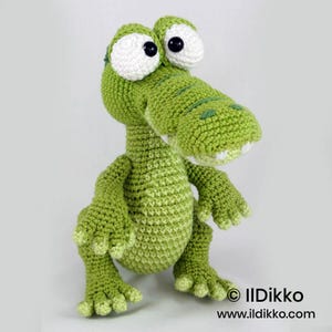 May include: A crochet stuffed animal shaped like a green crocodile with large white eyes and black pupils. The crocodile has a textured surface, a wide snout with visible white teeth, and four limbs with clawed feet. Text reads: &copy; IlDikko www.ildikko.com