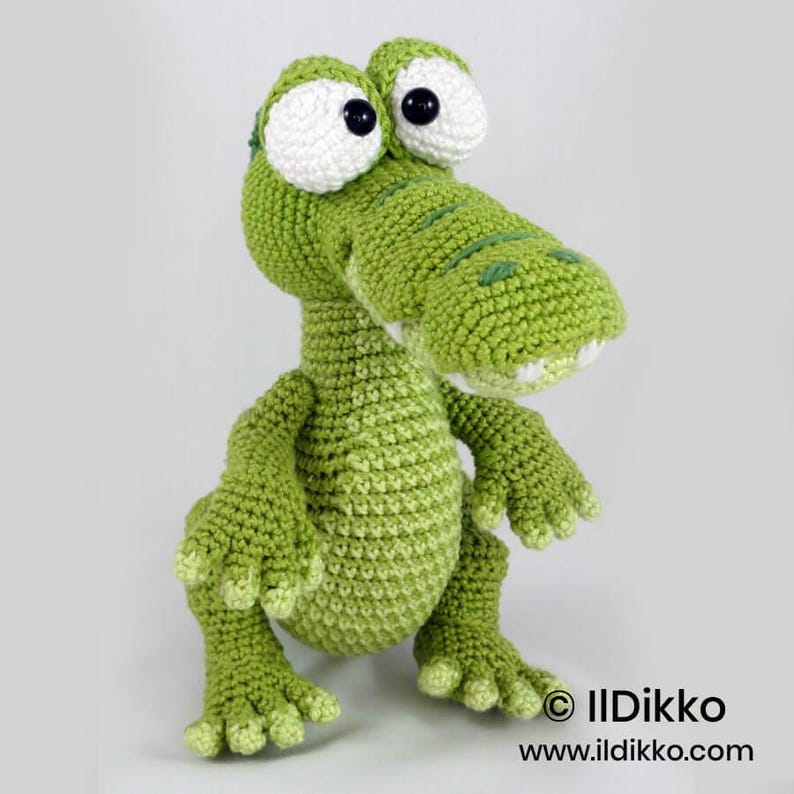May include: A crochet stuffed animal shaped like a green crocodile with large white eyes and black pupils. The crocodile has a textured surface, a wide snout with visible white teeth, and four limbs with clawed feet. Text reads: &copy; IlDikko www.ildikko.com