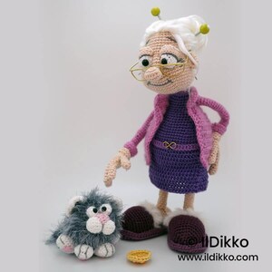 May include: A handmade crochet doll of an elderly woman wearing a purple dress and pink jacket, with glasses and white hair. A gray cat and a small yellow bowl are next to the doll. The doll is wearing burgundy slippers.