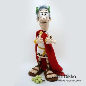 以下が含まれることがあります： 赤いトガとサンダルを履いたローマ皇帝の編みぐるみフィギュア。フィギュアは白いひげと緑色の月桂樹の輪を頭に付けています。フィギュアは両手を上げて親指を立てています。