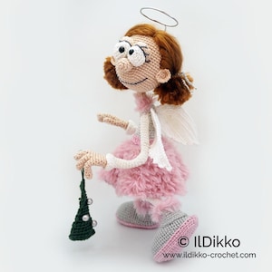 May include: A crocheted doll with auburn hair, a silver wire halo, and white wings. The doll wears a fluffy pink skirt, grey shoes with pink soles, and holds a small dark green triangular ornament with white beads. Text reads ILDikko.
