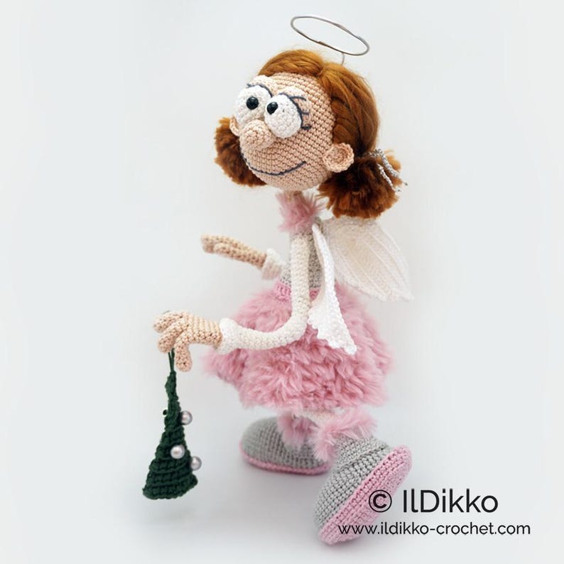 May include: A crocheted doll with auburn hair, a silver wire halo, and white wings. The doll wears a fluffy pink skirt, grey shoes with pink soles, and holds a small dark green triangular ornament with white beads. Text reads ILDikko.