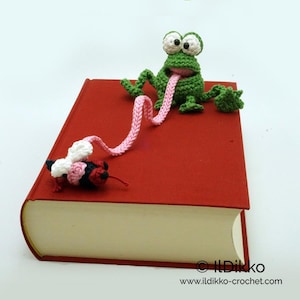 Puede incluir: Una rana verde de crochet con una lengua rosa que sobresale, sosteniendo un marcapáginas rosa de crochet. El marcapáginas está unido a una abeja roja de crochet con alas blancas. La rana y la abeja están encima de un libro rojo.