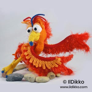 May include: A crocheted phoenix figurine featuring bright orange and red yarn with a textured, fuzzy appearance. The creature has large white eyes with black pupils, a yellow beak with a blue tip, and blue accents on its head and feet.