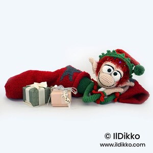 May include: A crocheted elf doll with red hair, a green hat, and a red stocking, accompanied by two small gift boxes. The elf has large white eyes and a beige face. The text "&copy; IlDikko www.ildikko.com" is printed in the bottom right corner.