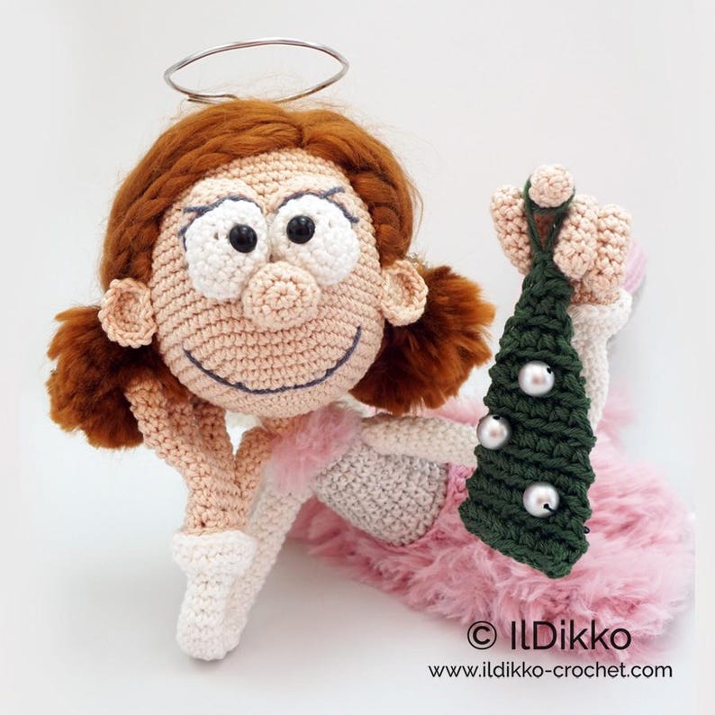 May include: A crocheted doll with auburn hair, a silver wire halo, and a pink fuzzy skirt. The doll has large black eyes and holds a small dark green crocheted Christmas tree decorated with three silver beads. Text reads: &copy; ILDikko www.ildikko-crochet.com