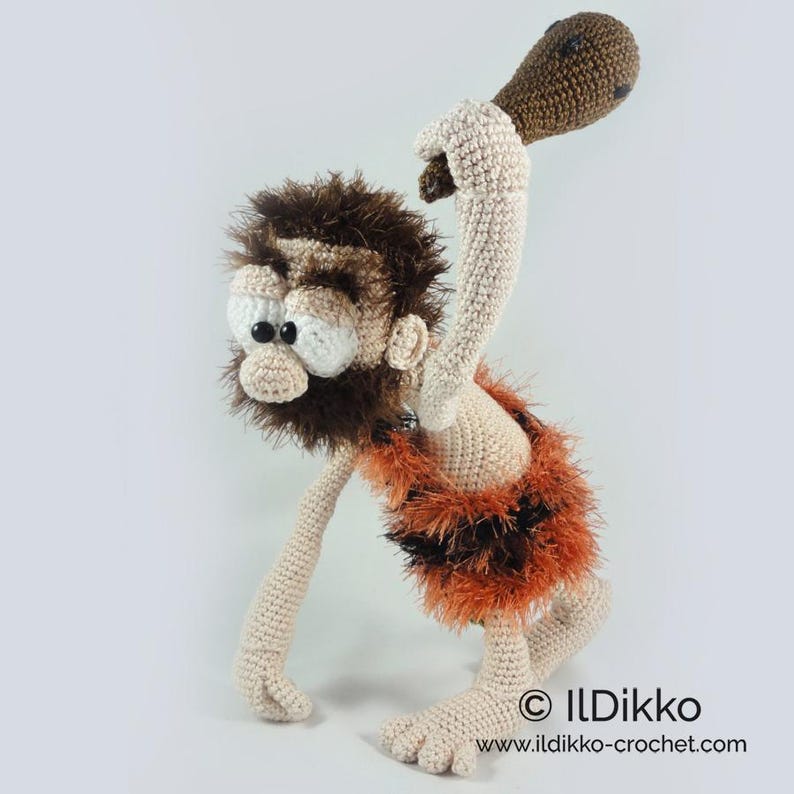 May include: A crocheted caveman doll with beige skin, a dark brown fuzzy beard and hair, and a skirt made of orange and black fuzzy yarn. The figure holds a brown crocheted club overhead and features large white eyes with black pupils. Text: ILDikko.