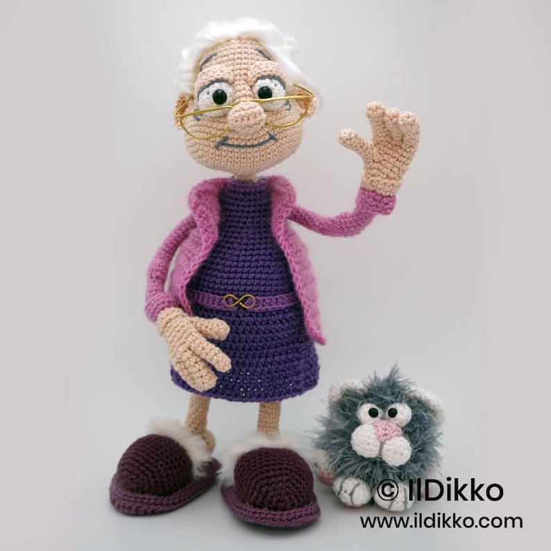 May include: A handmade crochet doll of an elderly woman wearing a purple dress and pink jacket, with glasses and a waving hand. The doll has matching slippers and is accompanied by a small, fluffy gray cat. The doll is a unique, handcrafted item.