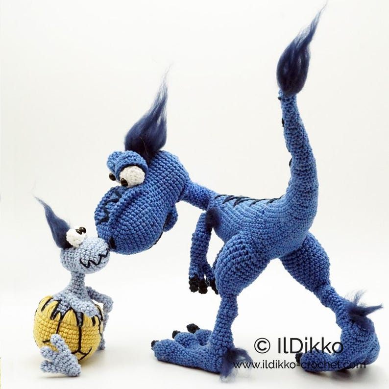 May include: Two crocheted figures: a large blue dinosaur with dark blue tufts and a smaller light blue creature emerging from a yellow egg with black markings. The text "&copy; ILDikko www.ildikko-crochet.com" is printed in the bottom right corner.