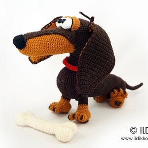 May include: A handmade crochet dachshund dog with a bone. The dog is brown and tan with a red collar and large, expressive eyes. The bone is off-white. The dog is sitting on a white surface.