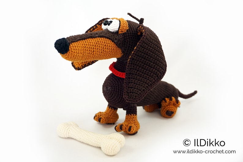 May include: A handmade crochet dachshund dog with a bone. The dog is brown and tan with a red collar and large, expressive eyes. The bone is off-white. The dog is sitting on a white surface.