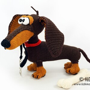May include: A handmade crochet dachshund dog with brown and tan yarn. The dog has large, floppy ears, a red collar, and a black leash. A cream-colored bone is next to the dog. The website address is visible on the bone.