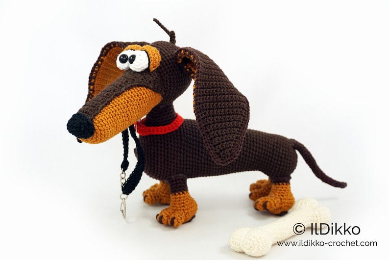 May include: A handmade crochet dachshund dog with brown and tan yarn. The dog has large, floppy ears, a red collar, and a black leash. A cream-colored bone is next to the dog. The website address is visible on the bone.