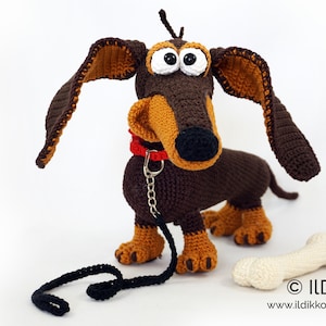 May include: Crocheted dachshund dog with large, floppy ears, a red collar, and a black leash. The dog is brown and tan with large, googly eyes. A crocheted bone is next to the dog. The dog is approximately 12 inches long.