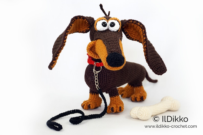 May include: Crocheted dachshund dog with large, floppy ears, a red collar, and a black leash. The dog is brown and tan with large, googly eyes. A crocheted bone is next to the dog. The dog is approximately 12 inches long.
