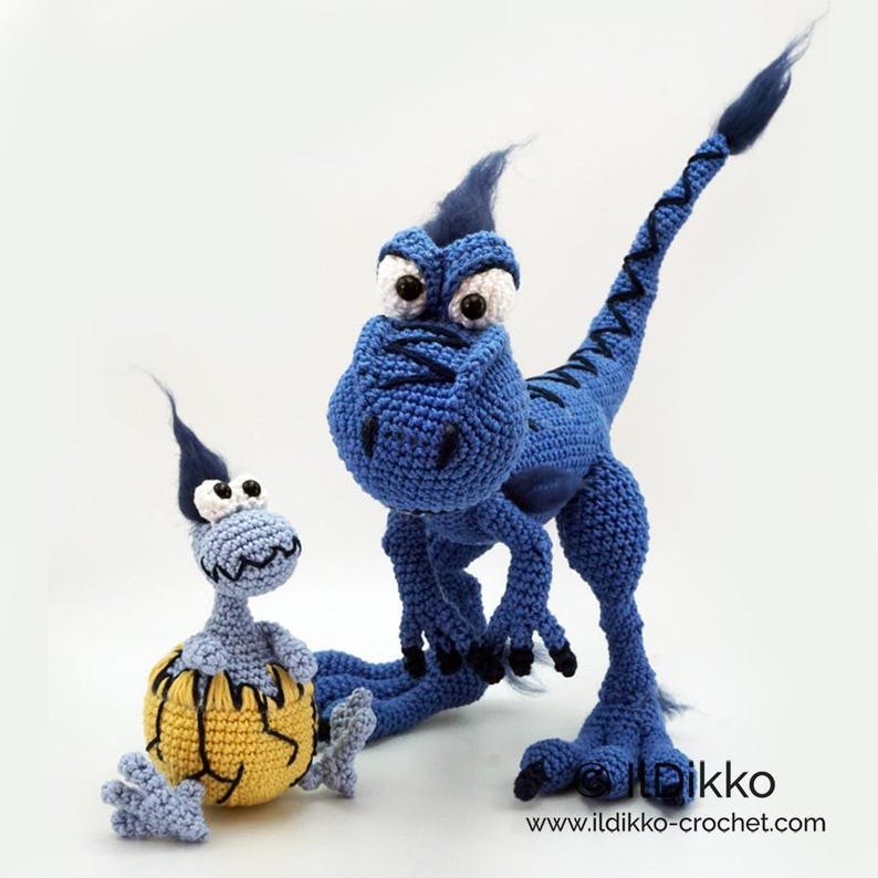 May include: Two crocheted dinosaur figures. The larger is dark blue with a long tail, crest, and black claws. The smaller is light blue, emerging from a yellow, cracked eggshell. Both have large white eyes with black pupils and text: Ildikko www.ildikko-crochet.com