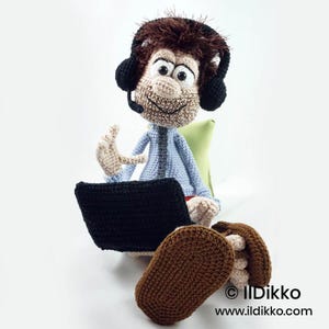 May include: A crocheted doll with brown hair, wearing black headphones with a microphone, a light blue shirt, and dark brown shoes. The doll is sitting and holding a black crocheted laptop. The text IlDikko www.ildikko.com is visible.