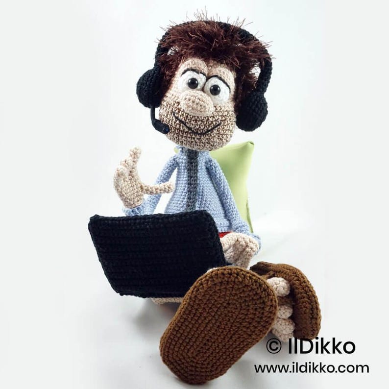 May include: A crocheted doll with brown hair, wearing black headphones with a microphone, a light blue shirt, and dark brown shoes. The doll is sitting and holding a black crocheted laptop. The text IlDikko www.ildikko.com is visible.