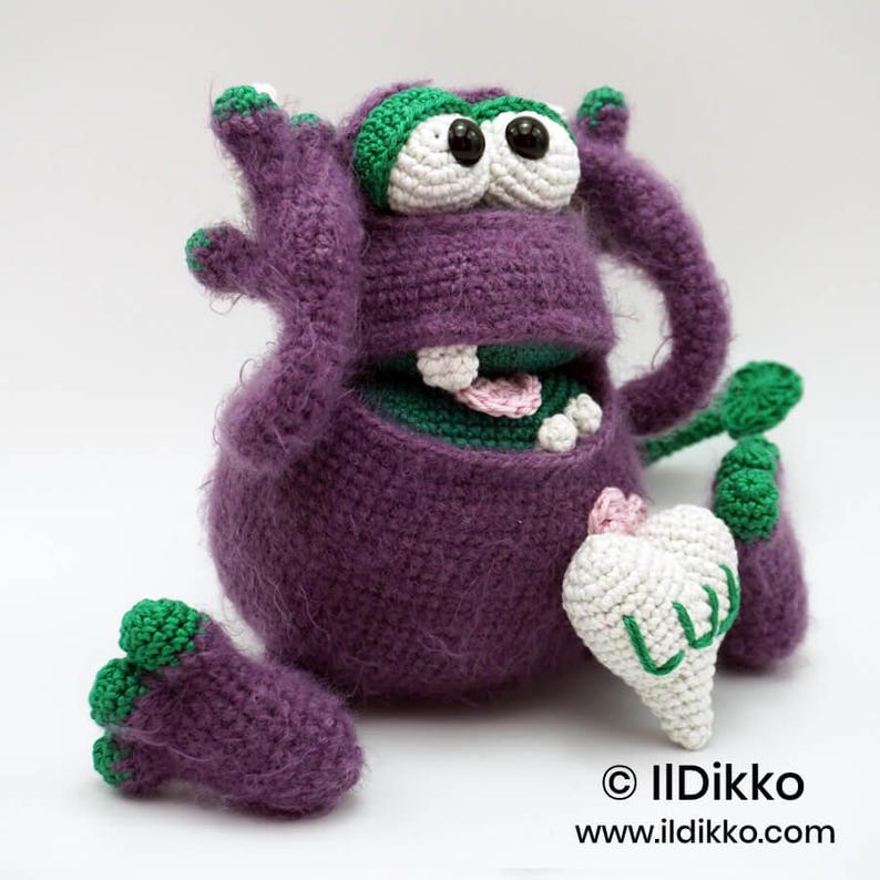 Puede incluir: Un monstruo de ganchillo morado con detalles verdes, grandes ojos blancos con pupilas negras y una boca ancha que muestra dientes y una lengua rosa. Sostiene un objeto blanco en forma de coraz&oacute;n con el texto verde LUV. El texto dice Ildikko.