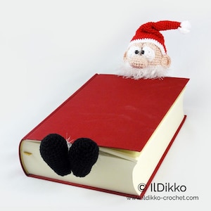 May include: A crochet bookmark featuring a Santa Claus head with a red and white hat and a white beard, paired with two black crochet boots. The figure is positioned on a red hardcover book. The text "IlDikko" and "ildikko-crochet.com" is visible.