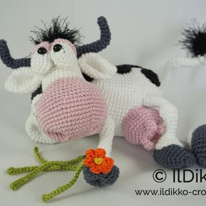 May include: A crocheted plush toy cow with black and white spots, pink udder, and a black tail. The cow is holding a crocheted orange flower.