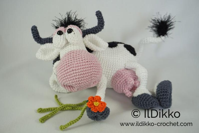 May include: A crocheted plush toy cow with black and white spots, pink udder, and a black tail. The cow is holding a crocheted orange flower.