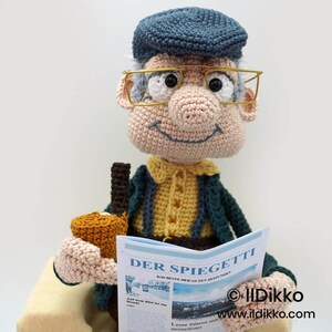 May include: A crocheted doll of an elderly man wearing a blue cap and gold-rimmed glasses, holding a newspaper and a brown bottle. The newspaper reads "DER SPIEGETTI". The doll is dressed in a yellow shirt and a dark green jacket.
