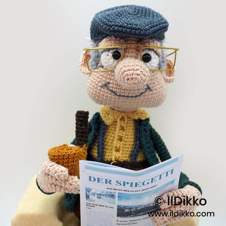 May include: A crocheted doll of an elderly man wearing a blue cap and gold-rimmed glasses, holding a newspaper and a brown bottle. The newspaper reads "DER SPIEGETTI". The doll is dressed in a yellow shirt and a dark green jacket.