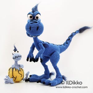 May include: A large blue crocheted dinosaur with black claws and a tuft of hair, standing next to a smaller light blue crocheted dinosaur emerging from a yellow cracked egg. The text ILDikko and www.ildikko-crochet.com is visible at the bottom.