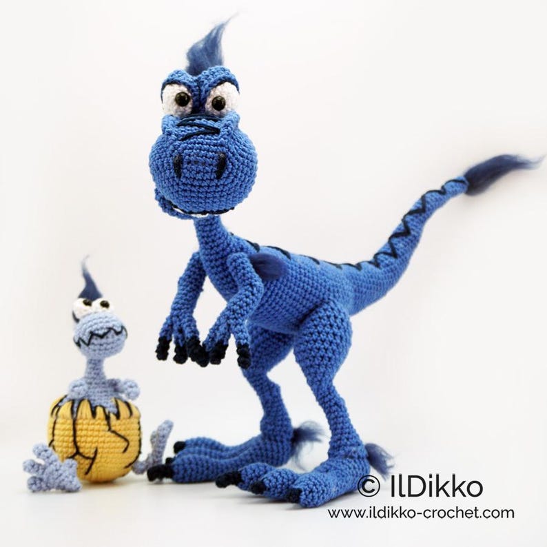 May include: A large blue crocheted dinosaur with black claws and a tuft of hair, standing next to a smaller light blue crocheted dinosaur emerging from a yellow cracked egg. The text ILDikko and www.ildikko-crochet.com is visible at the bottom.