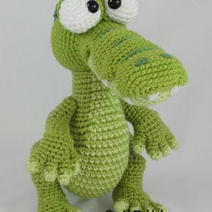May include: A green crocheted alligator toy with large white eyes and black pupils. The creature has a textured surface, a wide snout with visible white teeth, and four clawed feet. The text ILDikko and www.ildikko-crochet.com is visible.