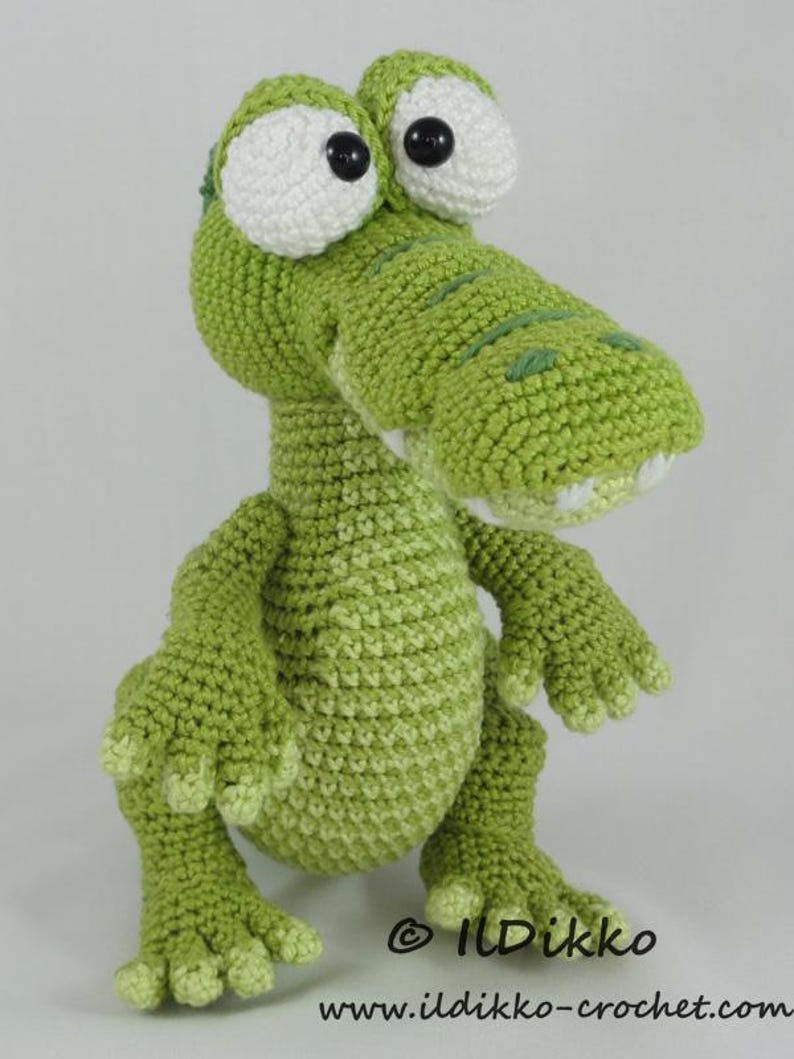 May include: A green crocheted alligator toy with large white eyes and black pupils. The creature has a textured surface, a wide snout with visible white teeth, and four clawed feet. The text ILDikko and www.ildikko-crochet.com is visible.