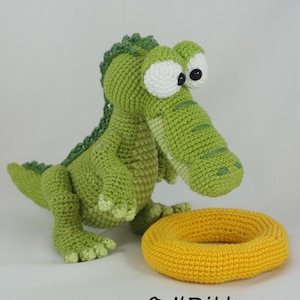 May include: A green crocheted crocodile toy with large white eyes and black pupils, featuring a textured back ridge. Beside the crocodile sits a yellow crocheted ring. The text ILDikko and www.ildikko-crochet.com is printed at the bottom.