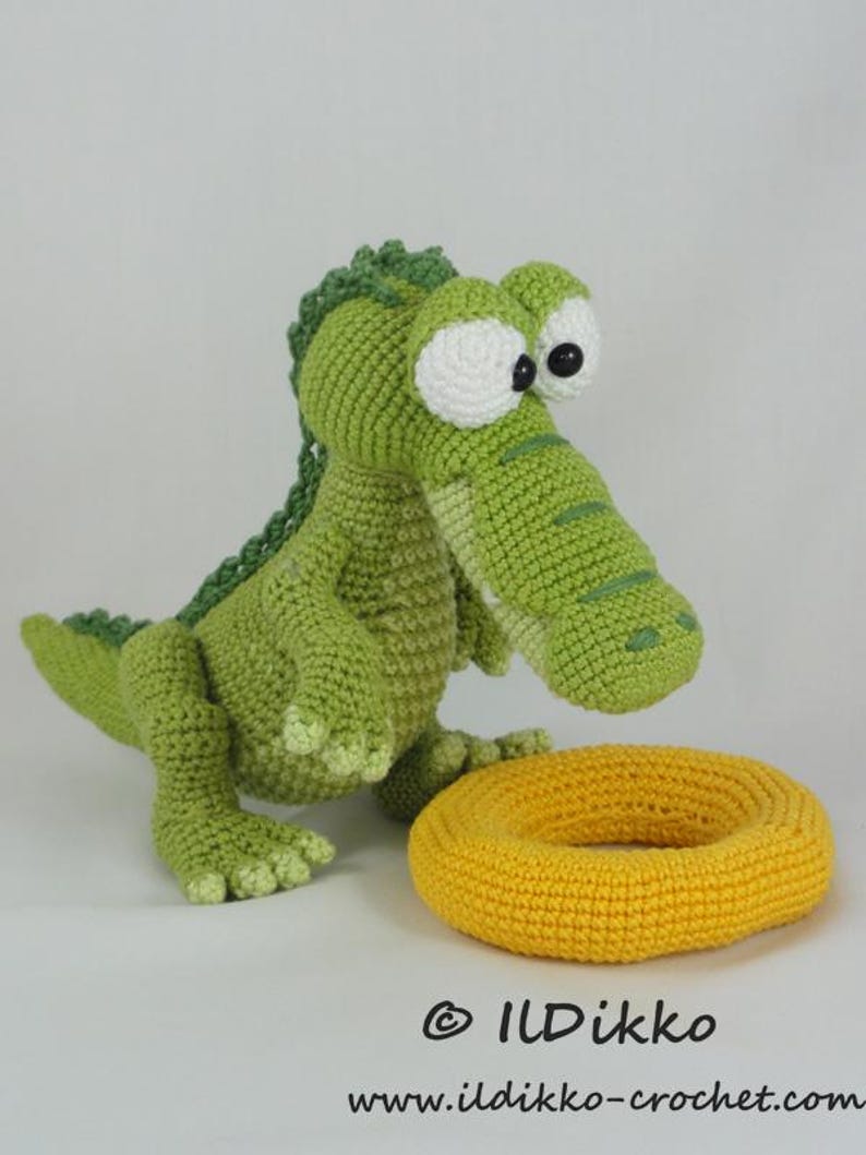 May include: A green crocheted crocodile toy with large white eyes and black pupils, featuring a textured back ridge. Beside the crocodile sits a yellow crocheted ring. The text ILDikko and www.ildikko-crochet.com is printed at the bottom.