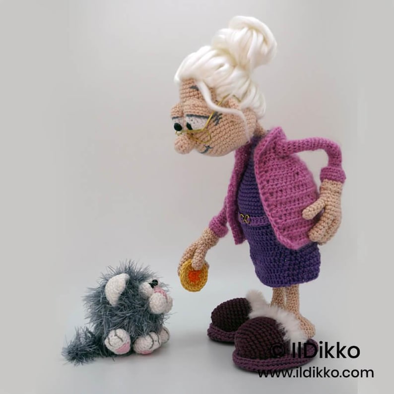 May include: Crocheted doll of a woman with white hair, glasses, and a purple outfit, interacting with a small gray crocheted cat. The woman is holding a small, round object. The cat has pink accents. The doll is handmade.