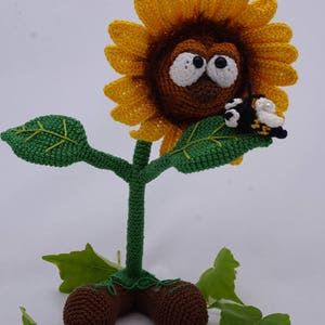 May include: A crocheted sunflower with a brown face and large eyes, wearing a yellow flower. The sunflower has a green stem and leaves, and is standing on brown feet. A small crocheted bee is perched on one of the leaves.