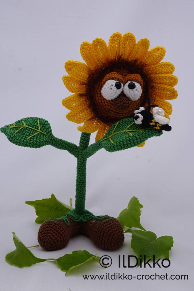 May include: A crocheted sunflower with a brown face and large eyes, wearing a yellow flower. The sunflower has a green stem and leaves, and is standing on brown feet. A small crocheted bee is perched on one of the leaves.