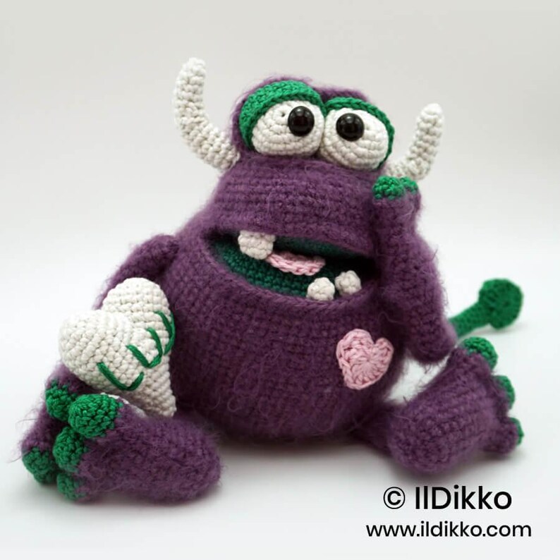 Puede incluir: Un monstruo de ganchillo morado con dos cuernos blancos, ojos grandes blancos con pupilas negras y p&aacute;rpados verdes. Tiene una boca ancha con dientes blancos, una lengua rosa y un peque&ntilde;o coraz&oacute;n rosa en el pecho. Sostiene un objeto en forma de coraz&oacute;n.