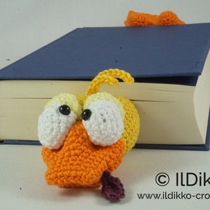 May include: A crocheted yellow and orange bird bookmark with large white eyes and a purple tongue sticking out. The bookmark is peeking out from behind a blue book.