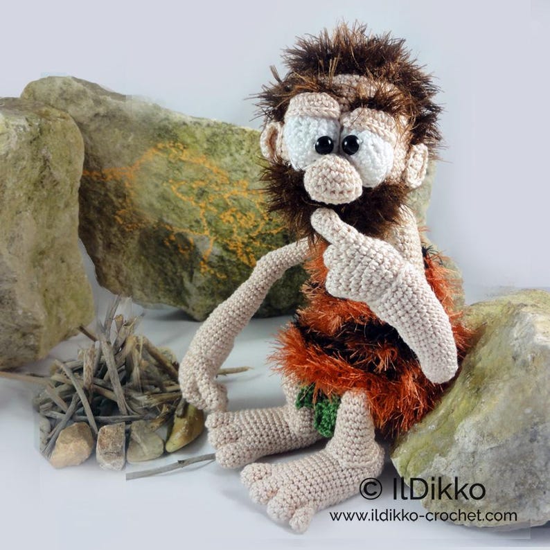 May include: A crocheted caveman doll with dark brown fuzzy hair and beard, large white eyes, and a beige skin tone. The figure wears a textured orange and brown garment with a small green leaf detail. Text reads: IlDikko www.ildikko-crochet.com.