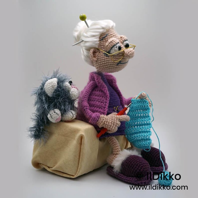 May include: A crocheted doll of an elderly woman with white hair, glasses, and a purple outfit, knitting a blue piece of fabric. A gray crocheted cat sits beside her. The doll is wearing purple slippers with white fur trim.