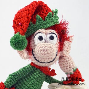 May include: A crocheted elf doll with beige skin, large white eyes, and red hair. The elf wears a red and green hat with a green pom-pom, a green shirt, and red cuffs. The text "© Ildikko www.ildikko.com" is printed in the bottom right corner.