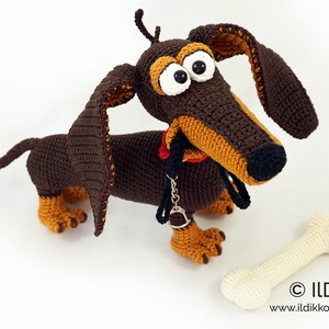 May include: A handmade crochet dachshund dog with large eyes, long ears, and a red collar. The dog is brown and tan, with a bone-shaped toy beside it. The image includes the text "&copy; ILDikko" and a website address.