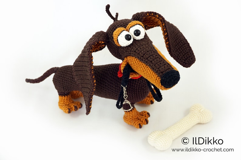 May include: A handmade crochet dachshund dog with large eyes, long ears, and a red collar. The dog is brown and tan, with a bone-shaped toy beside it. The image includes the text "&copy; ILDikko" and a website address.