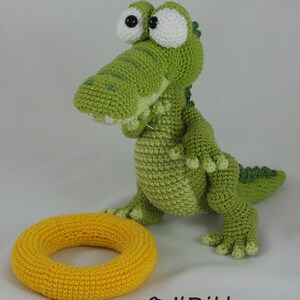May include: A green crocheted crocodile toy with large white eyes and black pupils, standing next to a yellow crocheted ring. The text "&copy; ILDikko" and "www.ildikko-crochet.com" is printed in black at the bottom of the image.