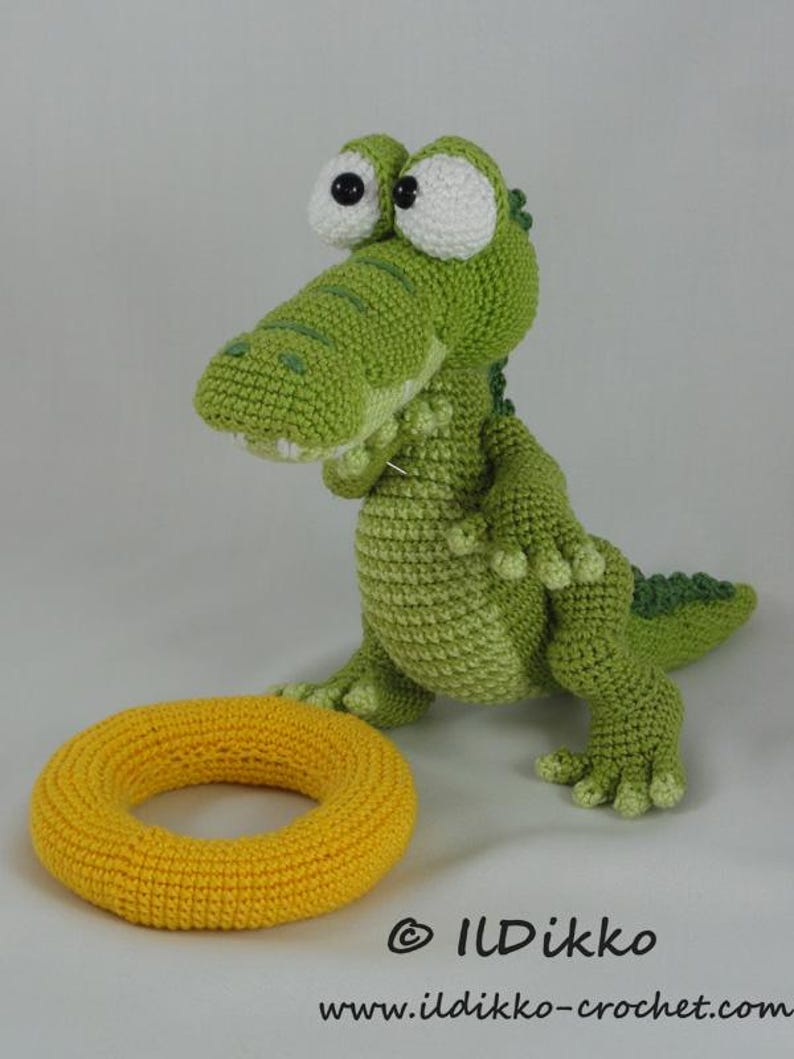 May include: A green crocheted crocodile toy with large white eyes and black pupils, standing next to a yellow crocheted ring. The text "&copy; ILDikko" and "www.ildikko-crochet.com" is printed in black at the bottom of the image.